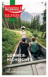 Zwei Personen sitzen auf einer Sommerrodelbahn im Grünen. Im Hintergrund sind hohe Bäume und Berge sichtbar.