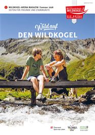 Zwei Personen sitzen am Ufer eines Bergsees und genießen die Natur. Im Hintergrund sind grüne Berge und ein blauer Himmel zu sehen.