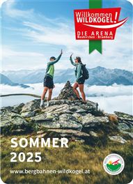 Zwei Personen stehen auf einem Berggipfel und jubeln. Im Hintergrund sind beeindruckende Berge und ein klarer Himmel zu sehen.