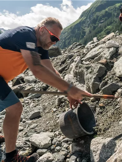 Een man werkt buiten en schept aarde in een emmer. Op de achtergrond zijn groene bergen en een heldere lucht zichtbaar.