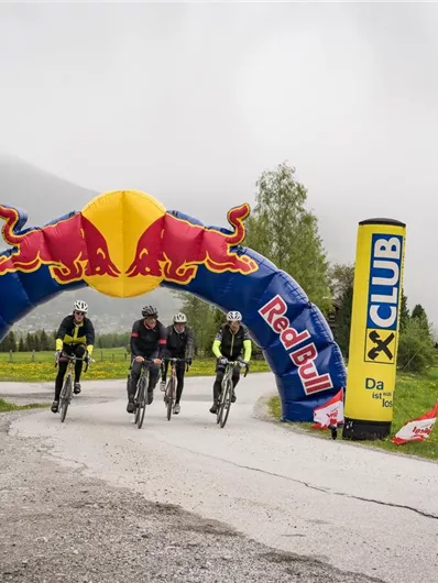Drei Radfahrer fahren unter einem Roten-Bull-Bogen hindurch. Im Hintergrund sind Nebel und eine große Bierflasche zu sehen.