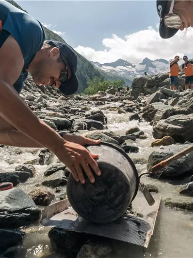 Two people are working in the high mountains and moving a large drum over rocks. In the background, there are people and an impressive mountain landscape.