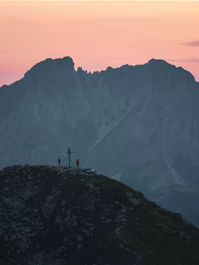 Ein imposanter Berg bei Sonnenuntergang, umgeben von sanften Hügeln. Silhouetten von Menschen stehen an der Spitze und genießen die Aussicht.