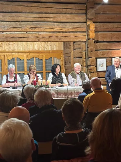 Eine Diskussionsrunde mit mehreren Personen sitzt auf einer Bühne in einem rustikalen Holzraum. Das Publikum besteht aus vielen Menschen, die aufmerksam zuhören.