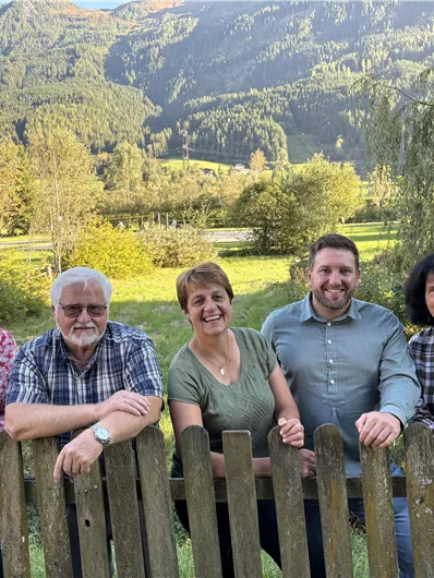 Eine Gruppe von fünf Personen steht an einem Holzzaun vor einer malerischen Landschaft. Im Hintergrund sind Berge und grüne Wiesen zu sehen.