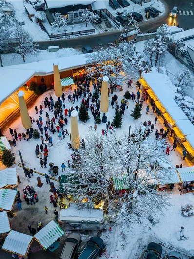 Ein winterlicher Weihnachtsmarkt mit vielen Besuchern in einem verschneiten Dorf. Lichter und Stände schaffen eine festliche Atmosphäre.