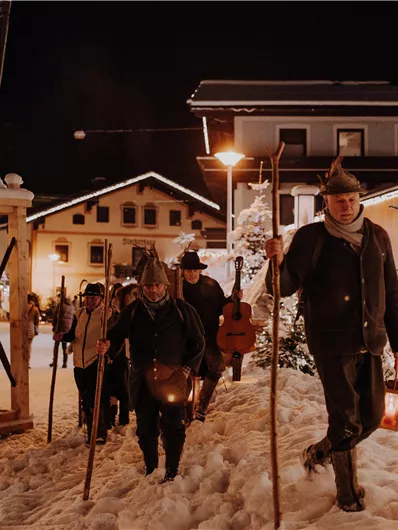 Eine winterliche Szene mit Menschen, die in warmen Kleidern durch den Schnee gehen. Die Umgebung ist festlich beleuchtet und es gibt historische Gebäude im Hintergrund.