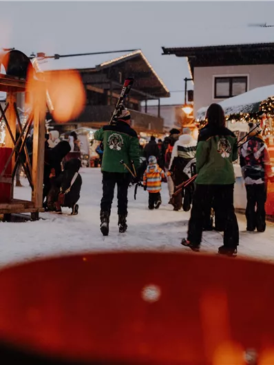 Ein winterlicher Weihnachtsmarkt mit Schneebedecktem Boden und festlich beleuchteten Ständen. Menschen schlendern vorbei und genießen die Atmosphäre.