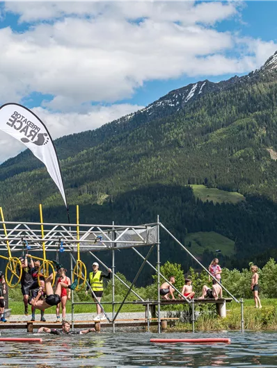 A group of people is standing on a platform by the water, surrounded by mountains. In the background, a beautiful landscape with green hills and a blue sky can be seen.