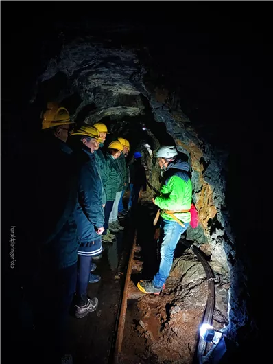 Eine Gruppe von Personen steht in einem dunklen Tunnel und trägt Helme. Ein Führer erklärt etwas, während sie sich um eine Schiene versammeln.