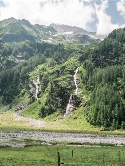 Eine malerische Berglandschaft mit Wasserfällen und dichtem grünen Wald. Der Himmel ist teilweise bewölkt und die Natur wirkt unberührt.