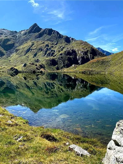 Ein malerischer Bergsee, umgeben von grünen Wiesen und majestätischen Bergen. Der klare Himmel spiegelt sich perfekt im ruhigen Wasser.