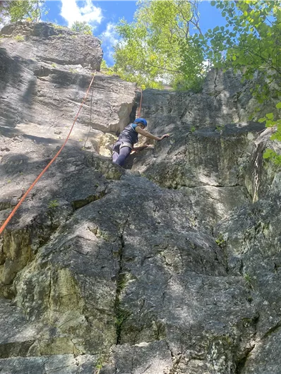 A person is climbing a rock face with a rope belay. In the background, trees and a blue sky are visible.