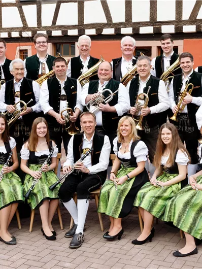 A brass band stands in traditional clothing on a paved area. The musicians are holding various instruments in their hands and smiling at the camera.