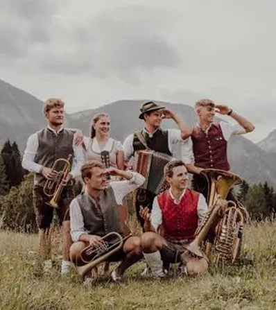 Eine Gruppe junger Musiker in traditioneller Tracht steht im Freien. Sie halten verschiedene Blasinstrumente und genießen die natürliche Umgebung mit Bergen im Hintergrund.