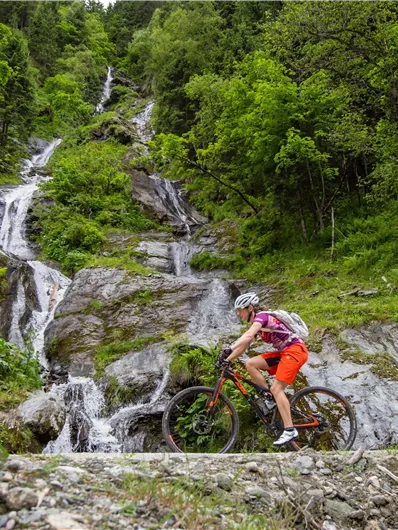 Two mountain bikers ride along a rocky path, surrounded by lush greenery and a waterfall in the background. The scene showcases an idyllic natural landscape in the mountains.