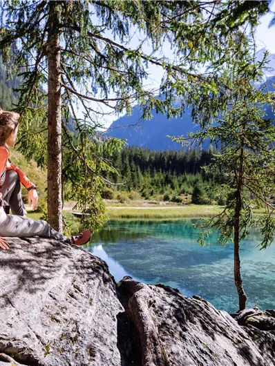 A person is sitting on a rock at the edge of a clear lake. In the background, high mountains and green trees can be seen.