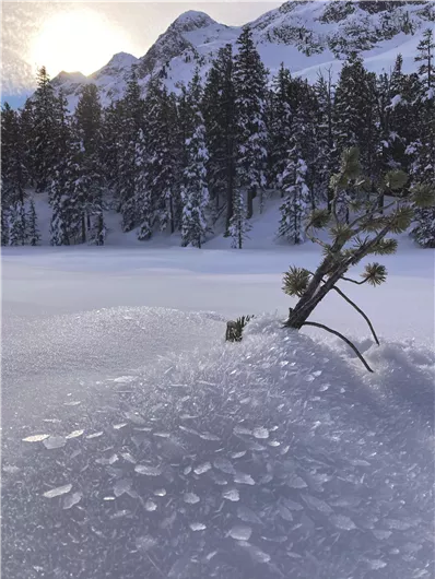 Eine schneebedeckte Landschaft mit Bäumen im Hintergrund. Die Sonne scheint über die Berge und einen ruhigen, gefrorenen See.