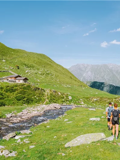 A green valley with a small cabin in the background. Two hikers walk along a clear stream with mountains in the background.