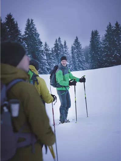 Eine Gruppe von Wanderern steht im Schnee, umgeben von schneebedeckten Bäumen. Einer der Wanderer trägt grüne Kleidung und hält Skistöcke in der Hand.