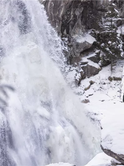 Ein Wanderer steht vor einem gefrorenen Wasserfall in einer winterlichen Landschaft. Die Umgebung ist schneebedeckt und vermittelt eine ruhige, idyllische Stimmung.