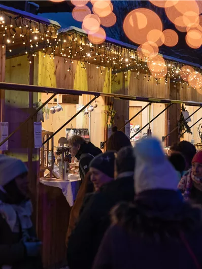 Ein festlicher Markt mit Holzständen, die mit Lichtern geschmückt sind. Menschen stehen zusammen und genießen die Atmosphäre.