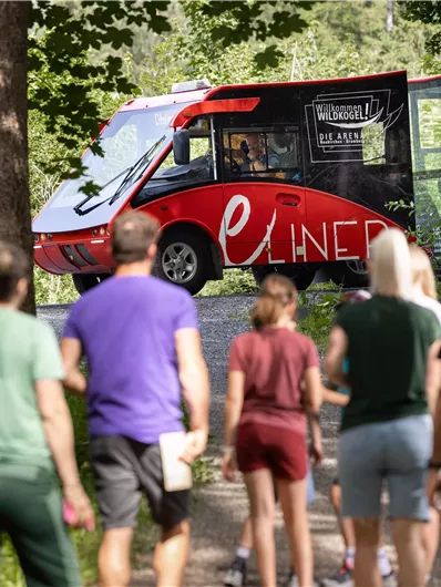 A group of people is walking along a woodland path while a red vehicle with the inscription "e-LINER" stands in the background. The scenery is green and inviting, surrounded by trees.