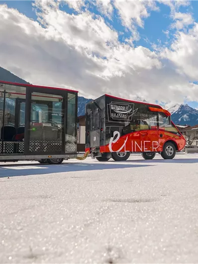 A red electric vehicle with a trailer is standing on a snowy surface. In the background, mountains and a blue sky with clouds can be seen.