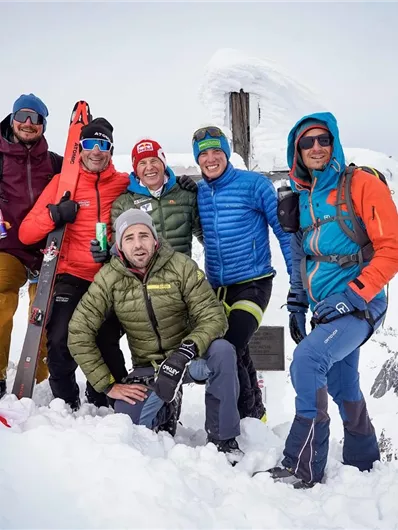 Eine Gruppe von sechs Personen steht auf einem schneebedeckten Gipfel. Sie tragen Wintersportkleidung und posieren fröhlich vor einer markierten Bergspitze.