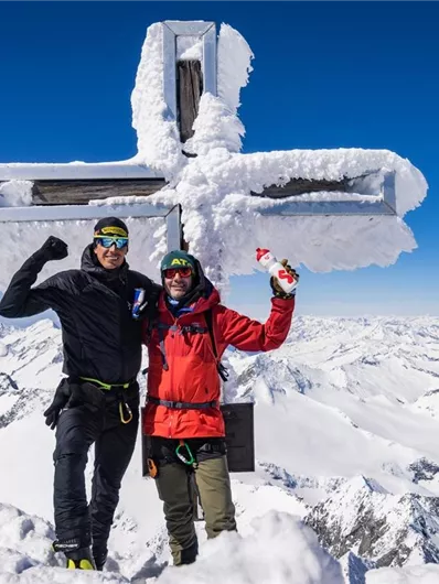Zwei glückliche Bergsteiger stehen an einem schneebedeckten Gipfel mit einem großen Kreuz. Im Hintergrund erstreckt sich eine beeindruckende Berglandschaft unter klarem blauen Himmel.