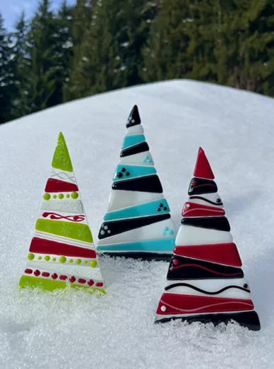 Drie decoratieve kerstbomen van glas staan in de sneeuw. Ze zijn in verschillende kleuren en patronen ontworpen en steken af tegen het groene bos.