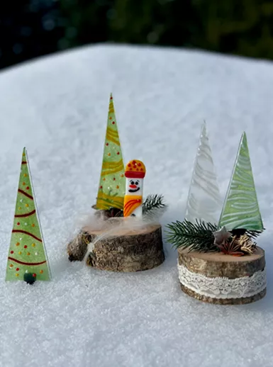 Kleurige kerstboomdecoraties van glas en hout staan op een sneeuwdeken. De decoraties bestaan uit gestileerde dennenbomen en een grappige kerstman.