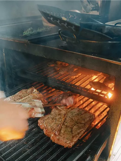 Een grill met sappig vlees dat op een gloeiende ondergrond wordt bereid. Rook en damp stijgen op terwijl een kok het vlees omdraait.