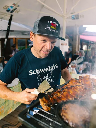 Een man is vlees aan het grillen en draagt een T-shirt met de tekst "Schweini's". Op de achtergrond zijn mensen en een grote luifel te zien.
