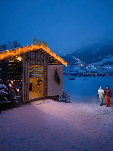 Eine gemütliche Hütte im Schnee, geschmückt mit Lichtern, in der Abenddämmerung. Zwei Personen spazieren in der frostigen Landschaft.