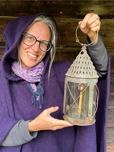 Eine Person in einem lila Umhang hält eine alte Laterne. Im Hintergrund sind Holzwände zu sehen.