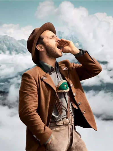 A man in traditional attire is calling out into the landscape. In the background, mountains and clouds can be seen.