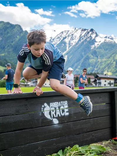A boy jumps over an obstacle in a picturesque landscape with mountains in the background. It is a sunny day with some clouds in the sky.