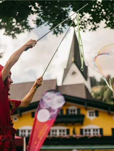 Ein Clown in roter Kleidung zaubert mit riesigen Seifenblasen. Im Hintergrund ist ein buntes Gebäude und Bäume zu sehen.
