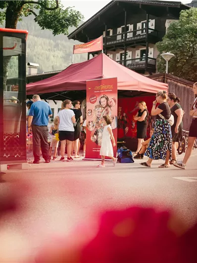 Eine belebte Straße mit einem roten Veranstaltungszelt. Menschen stehen zusammen und schauen neugierig zu einem Stand.