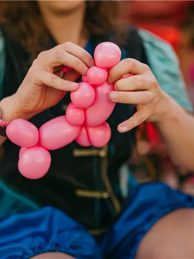 Eine Person formt mit rosa Luftballons eine Figur. Im Hintergrund sind Menschen und eine festliche Atmosphäre zu sehen.