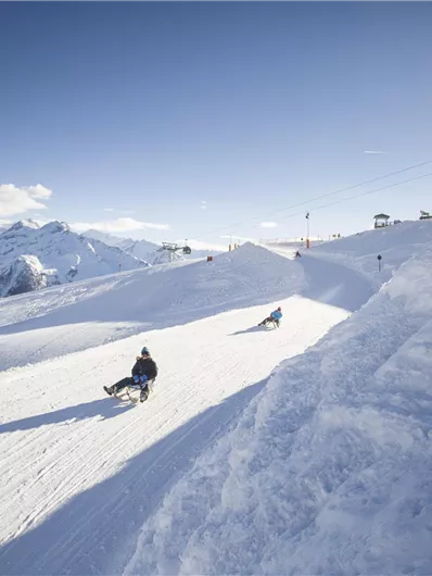 Eine beeindruckende Schneelandschaft mit Bergen und einer klaren, blauen Himmel. Zwei Schlittenfahrer gleiten die Piste hinunter.