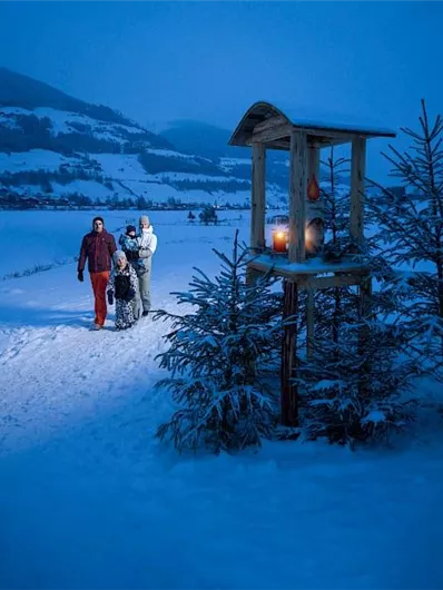 Eine winterliche Landschaft mit frischem Schnee und schneebedeckten Bäumen. Zwei Menschen gehen einen gemütlichen Weg entlang, während eine Laterne in der Nähe leuchtet.