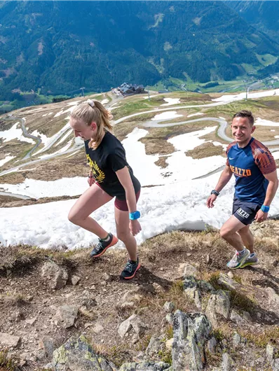 Two people are hiking up a steep mountain slope, surrounded by snowy terrain and a picturesque landscape. In the background, green valleys and winding roads can be seen.
