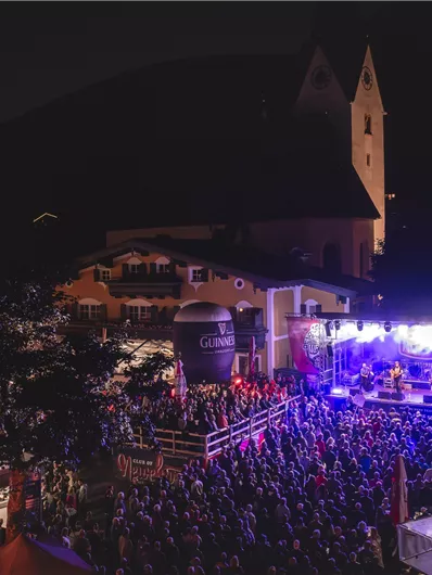 A lively concert under the night sky with a large crowd. Colorful lights illuminate the stage and the surroundings.