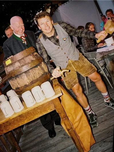 Eine fröhliche Szene mit einem Mann in traditioneller Kleidung, der ein Bierfass anzapft. Im Hintergrund sind weitere Gäste, die sich unterhalten und die Atmosphäre genießen.
