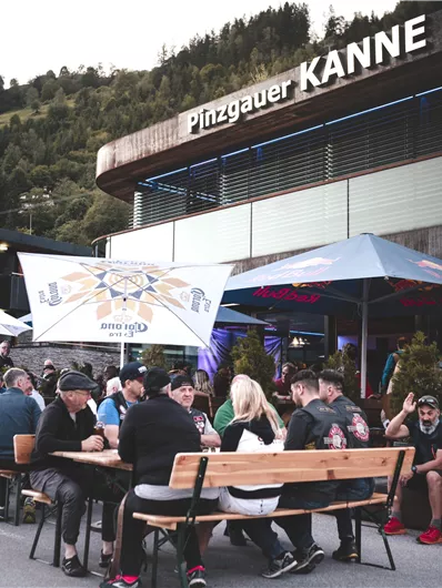 A lively terrace in front of a modern restaurant called "Pinzgauer Kanne". People are sitting together and enjoying drinks in a beautiful setting.