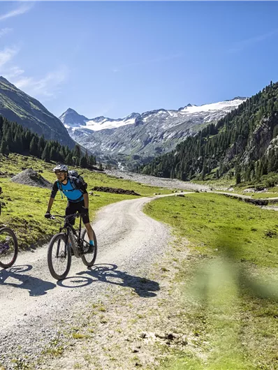 Two cyclists ride along a gravel path in the mountains. In the background, green valleys and majestic mountains stretch under a clear blue sky.