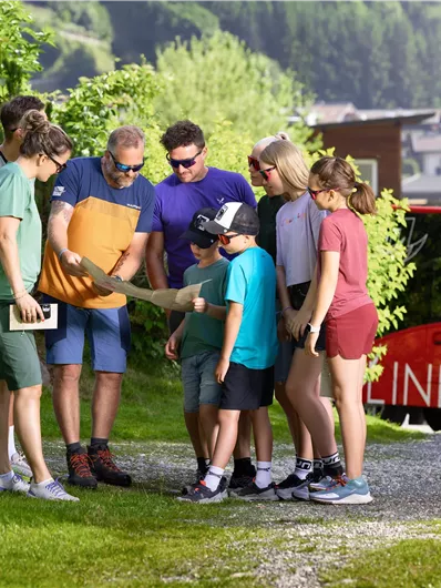 A group of people is looking at a map. In the background, green hills and a red vehicle are visible.