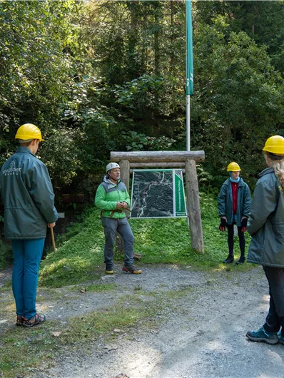 Eine Gruppe von sieben Personen trägt Helme und steht im Freien. Sie hören gespannt einem Mann zu, der an einem Schild spricht, umgeben von Bäumen.
